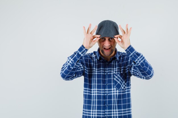 Joven sosteniendo su gorra en camisa y luciendo guapo. vista frontal.