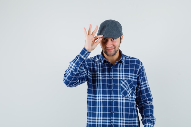 Joven sosteniendo su gorra en camisa y luciendo guapo. vista frontal.