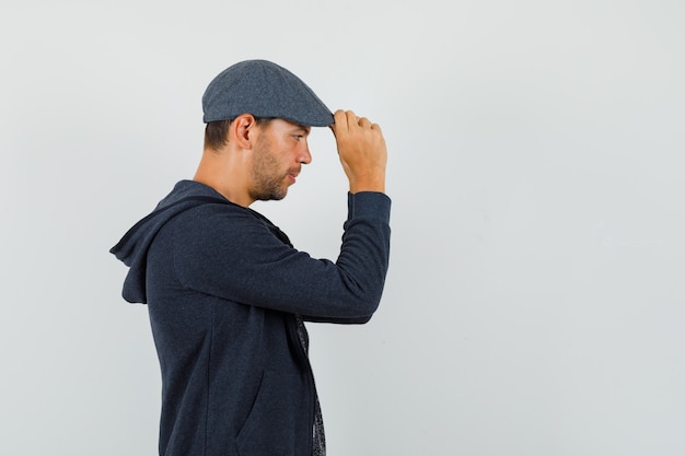 Joven sosteniendo su gorra en camiseta, chaqueta y mirando pensativo.