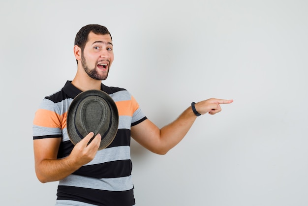 Joven sosteniendo su sombrero y apuntando hacia el lado en camiseta, vista frontal.