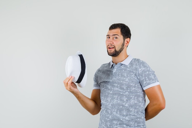 Joven sosteniendo su sombrero mientras posa con camiseta a rayas, sombrero y luciendo valiente. vista frontal.