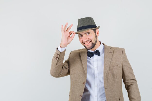 Joven sosteniendo su sombrero en traje y mirando alegre. vista frontal.