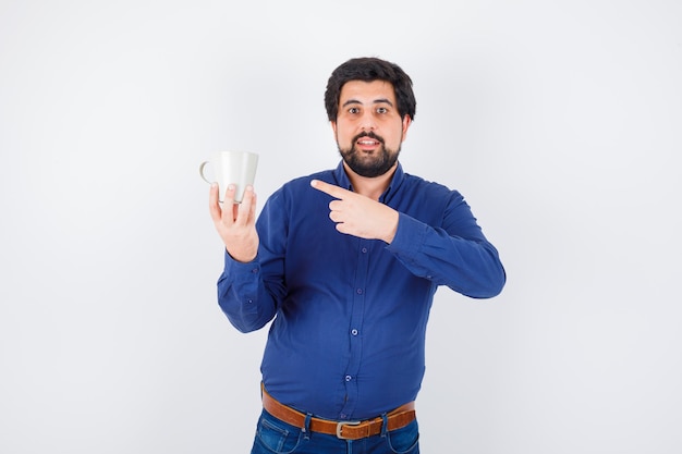 Joven sosteniendo la taza y apuntando hacia ella con camisa azul y jeans y luciendo optimista. vista frontal.