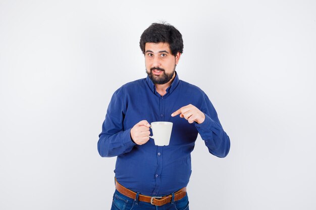 Joven sosteniendo la taza y apuntando a ella con camisa azul y jeans y mirando serio, vista frontal.