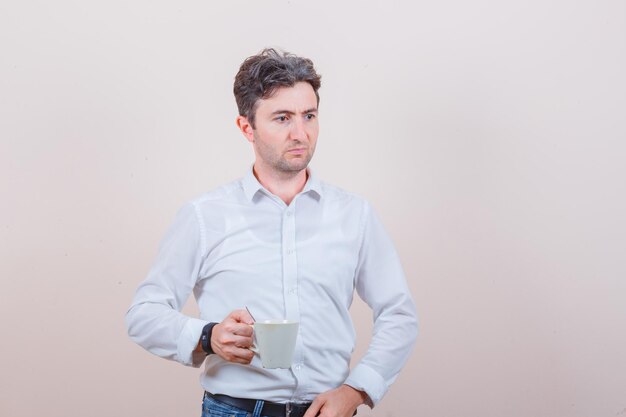 Joven sosteniendo una taza de bebida en camisa blanca, jeans y mirando pensativo