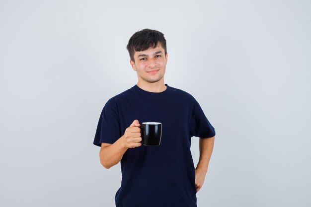 Joven sosteniendo una taza de bebida en camiseta negra y mirando orgulloso, vista frontal.