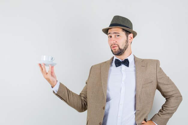 Joven sosteniendo una taza blanca con platillo en traje, sombrero y mirando confiado, vista frontal.