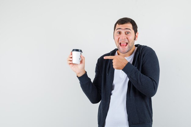 Joven sosteniendo una taza de café para llevar, apuntando con el dedo índice en una camiseta blanca y una sudadera con capucha negra con cremallera frontal y mirando sorprendido, vista frontal.