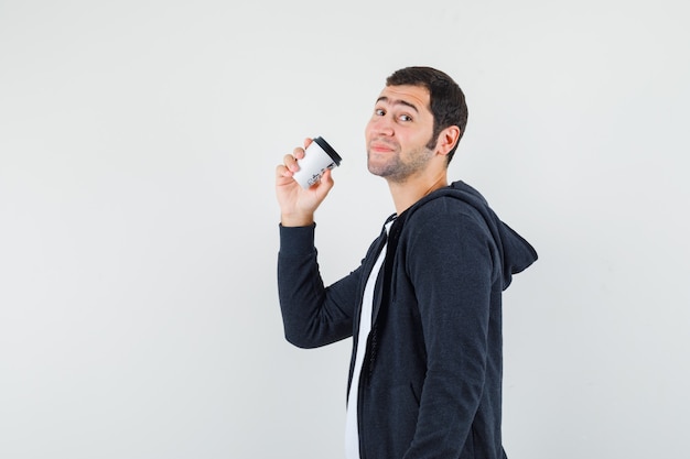 Joven sosteniendo una taza de café para llevar, sonriendo con camiseta blanca y sudadera con capucha negra con cremallera frontal y mirando feliz, vista frontal.