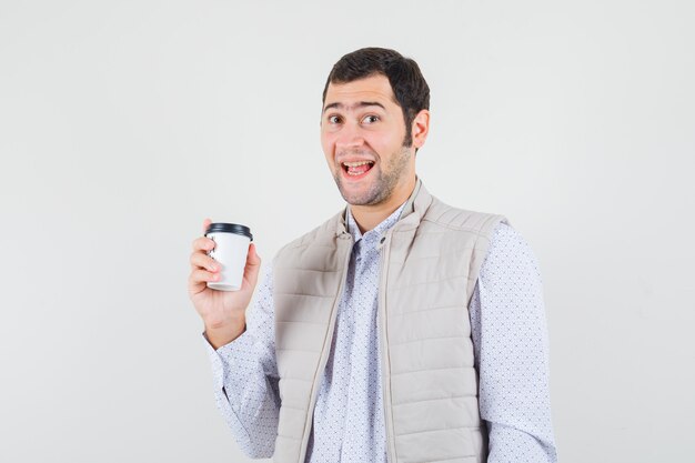 Joven sosteniendo una taza de café y riendo con chaqueta beige y gorra y mirando feliz. vista frontal.