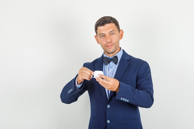 Joven sosteniendo una taza de café turco en traje y mirando contento.