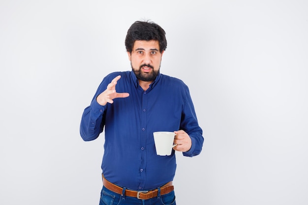 Joven sosteniendo la taza y estirando la mano como sosteniendo algo en camisa azul y jeans y mirando serio, vista frontal.