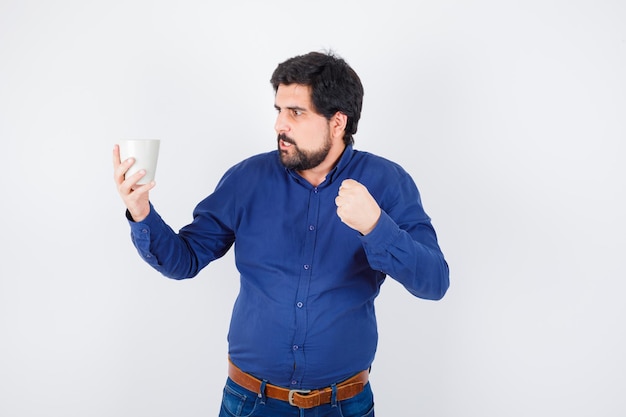 Joven sosteniendo la taza y mirándola, apretando el puño con camisa azul y jeans y mirando furioso. vista frontal.