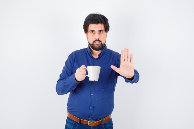 Joven sosteniendo la taza y mostrando la señal de pare en camisa azul y jeans y mirando serio, vista frontal.