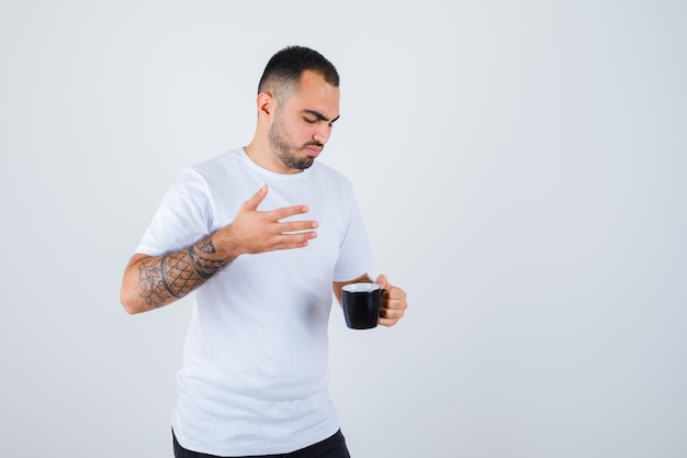 Joven sosteniendo una taza de té y estirando la mano derecha en camiseta blanca y pantalón negro y mirando enfocado