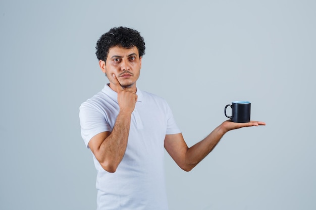 Joven sosteniendo una taza de té mientras se inclina la barbilla en la mano en camiseta blanca y jeans y mirando pensativo, vista frontal.