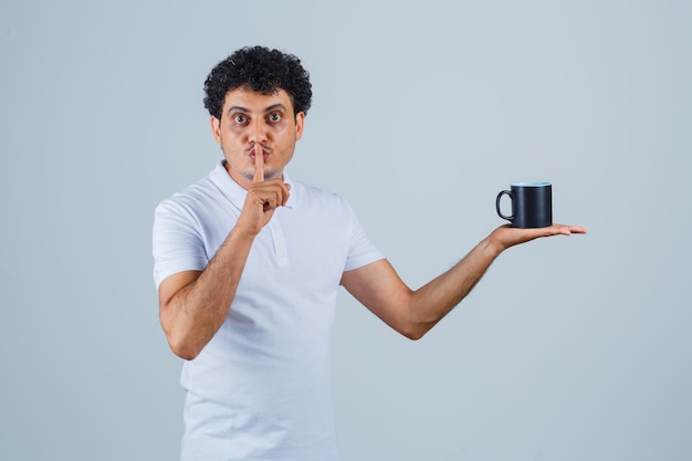 Joven sosteniendo una taza de té mientras muestra un gesto de silencio en camiseta blanca y jeans y parece serio. vista frontal.