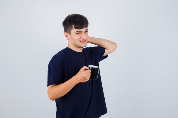 Joven sosteniendo una taza de té mientras se rasca la cabeza en una camiseta negra y mira olvidadiza, vista frontal.