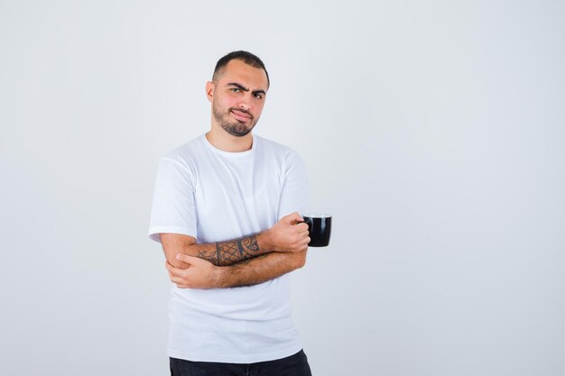 Joven sosteniendo una taza de té y de pie con los brazos cruzados en camiseta blanca y pantalón negro y mirando feliz