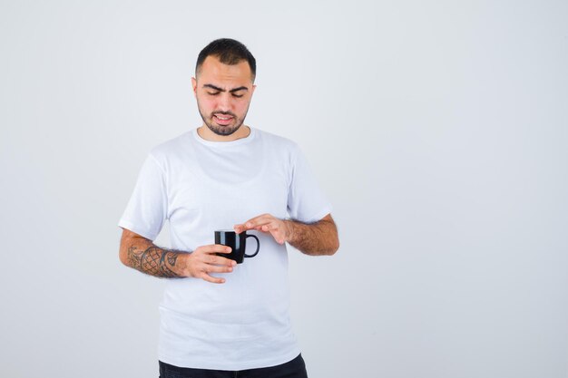 Joven sosteniendo una taza de té y poniendo la mano sobre ella en camiseta blanca y pantalón negro y mirando enfocado