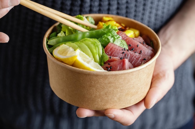 Joven sosteniendo un tazón de atún crudo saludable con verduras servidas en un tazón de papel