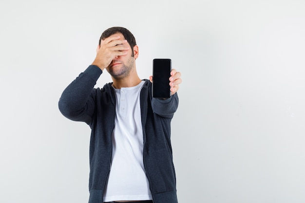 Joven sosteniendo el teléfono y cubriendo los ojos con una mano en una camiseta blanca y una sudadera con capucha negra con cremallera frontal y mirando molesto, vista frontal.