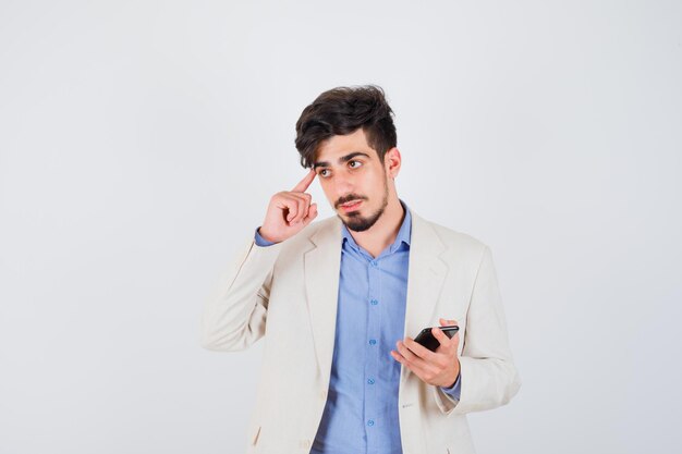 Joven sosteniendo el teléfono inteligente y de pie en pose de pensamiento en camiseta azul y chaqueta de traje blanco y mirando pensativo