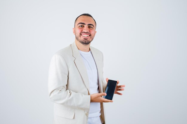 Joven sosteniendo teléfono móvil en camiseta blanca, chaqueta y mirando feliz, vista frontal.