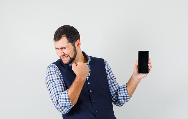 Joven sosteniendo el teléfono móvil, mostrando el gesto de ganador en camisa