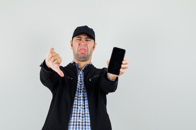 Joven sosteniendo teléfono móvil, mostrando el pulgar hacia abajo en camisa, chaqueta, gorra y mirando descontento.