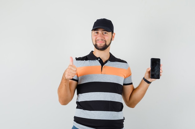 Joven sosteniendo el teléfono móvil, mostrando el pulgar hacia arriba en camiseta, gorra y mirando positivo, vista frontal.