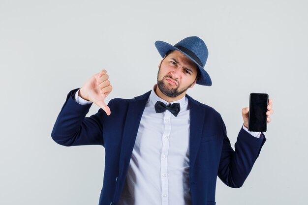 Joven sosteniendo el teléfono móvil con el pulgar hacia abajo en traje, sombrero y mirando insatisfecho, vista frontal.