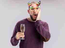 Foto gratuita joven en suéter morado y gafas divertidas sosteniendo una copa de champán mirando a la cámara asombrado y sorprendido de pie sobre fondo blanco.
