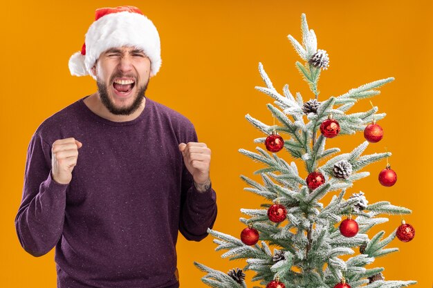 Joven en suéter morado y gorro de Papá Noel apretando los puños gritando con expresión agresiva de pie junto al árbol de Navidad sobre fondo naranja
