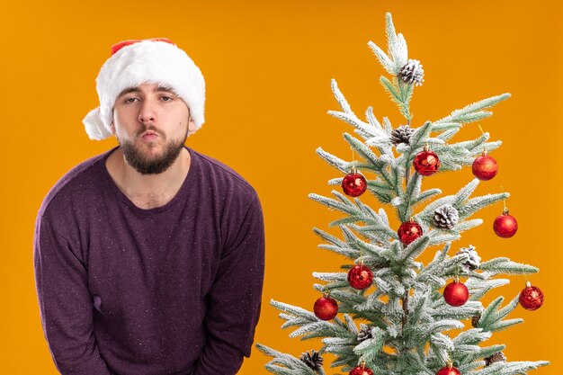 Joven en suéter morado y gorro de Papá Noel con aspecto cansado y aburrido de pie junto al árbol de Navidad sobre fondo naranja