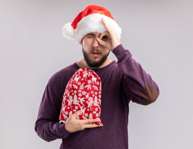Joven en suéter morado y gorro de Papá Noel con bolsa roja con regalos haciendo ok cantar mirando a través de este cartel de pie sobre fondo blanco