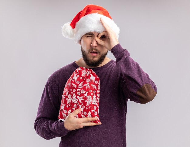 Joven en suéter morado y gorro de Papá Noel con bolsa roja con regalos haciendo ok cantar mirando a través de este cartel de pie sobre fondo blanco