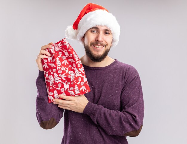 Joven en suéter morado y gorro de Papá Noel con bolsa roja con regalos mirando a la cámara con una sonrisa en la cara de pie sobre fondo blanco.