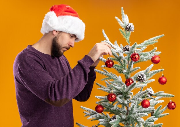 Joven en suéter morado y gorro de Papá Noel colgando juguetes en el árbol de navidad con cara seria de pie sobre fondo naranja