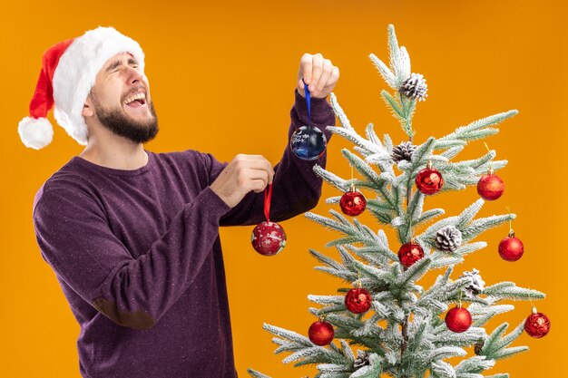 Joven en suéter morado y gorro de Papá Noel colgando juguetes en el árbol de Navidad