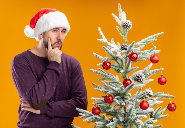 Joven en suéter morado y gorro de Papá Noel con expresión pensativa en la cara pensando de pie junto al árbol de Navidad sobre la pared naranja