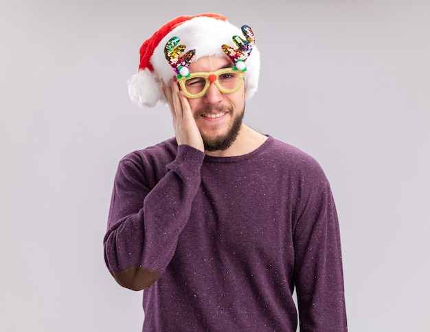 Joven en suéter morado y gorro de Papá Noel con gafas divertidas mirando a la cámara sonriendo feliz y positivo y guiñando un ojo sobre fondo blanco.