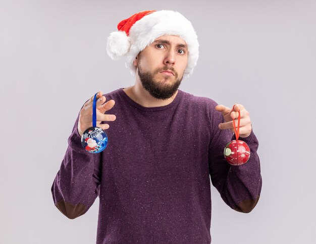 Joven en suéter morado y gorro de Papá Noel con gafas divertidas sosteniendo bolas de navidad mirando a la cámara confundido de pie sobre fondo blanco