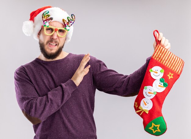 Joven en suéter morado y gorro de Papá Noel con gafas divertidas sosteniendo una media de Navidad confundido haciendo gesto de defensa con las manos de pie sobre la pared blanca