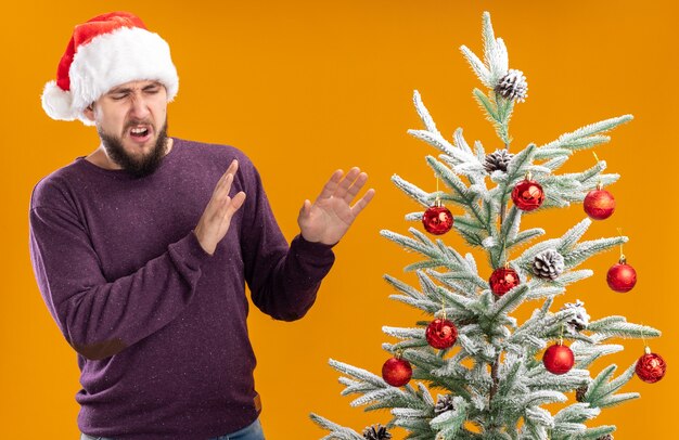 Joven en suéter morado y gorro de Papá Noel mirando el árbol de Navidad con expresión de disgusto haciendo gestos de defensa de pie sobre la pared naranja