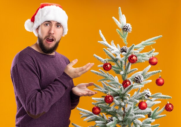 Joven en suéter morado y gorro de Papá Noel mirando asombrado y sorprendido de pie junto al árbol de Navidad sobre fondo naranja