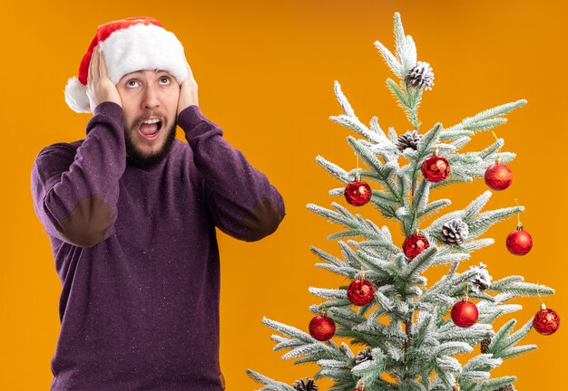 Joven en suéter morado y gorro de Papá Noel mirando asombrado y sorprendido de pie junto al árbol de Navidad sobre fondo naranja