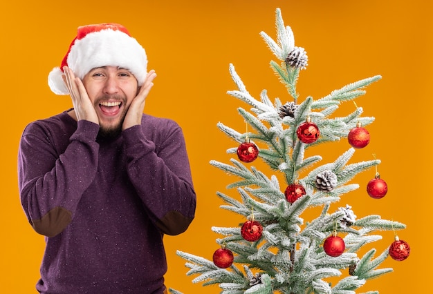 Foto gratuita joven en suéter morado y gorro de papá noel mirando a la cámara emocionado y loco feliz de pie junto al árbol de navidad sobre fondo naranja