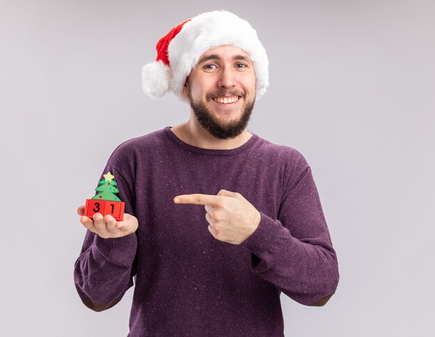 Joven en suéter morado y gorro de Papá Noel mostrando cubos con fecha de año nuevo apuntando con el dedo índice a él feliz y alegre de pie sobre fondo blanco.