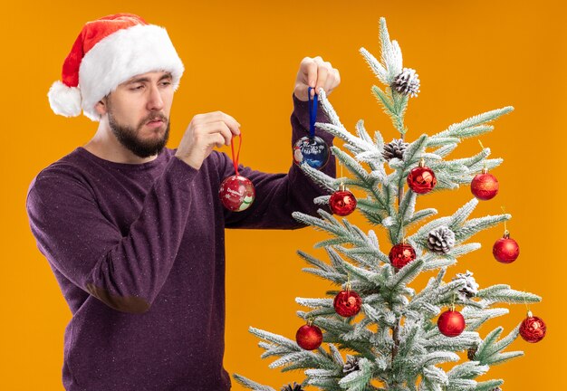 Joven en suéter morado y gorro de Papá Noel de pie junto al árbol de Navidad con juguetes mirando confundido